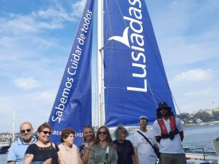 Fotografia de alguns Associados acompanhados pelo monitor antes de entrarem para o catamaran. Atrás do grupo vêem-se as velas do catamaran, de cor azul e a branco o seguinte texto "Sabemos Cuidar de Todos - Lusíadas".